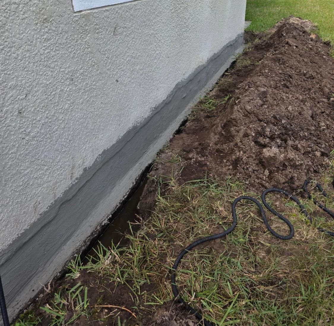 Grey waterproofing coat along the base of a stucco wall, with a trench dug beside it and the soil piled on the lawn Grey waterproofing coat along the base of a stucco wall, with a trench dug beside it and the soil piled on the lawn