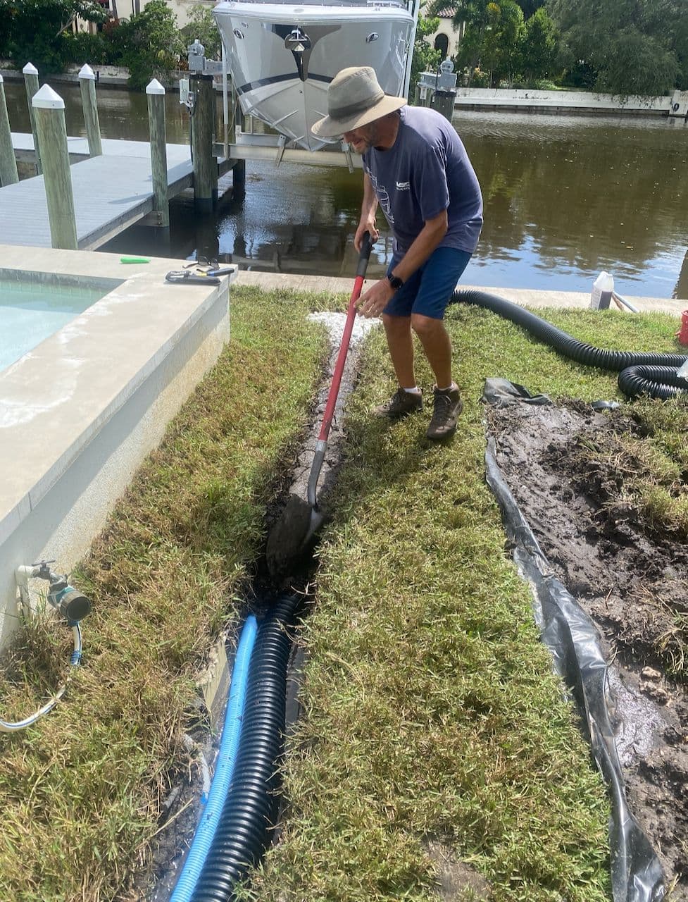 Worker digging a trench for a black corrugated French drain pipe along a grassy waterfront property next to a pool Worker digging a trench for a black corrugated French drain pipe along a grassy waterfront property next to a pool
