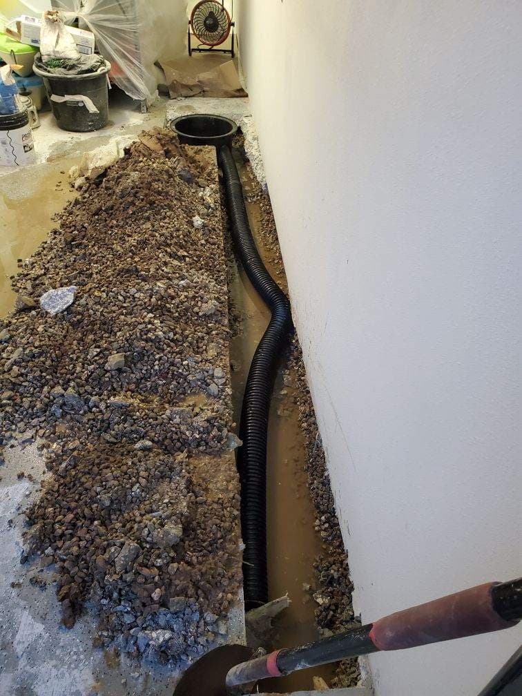 Interior basement trench along a wall showing a black corrugated drainage pipe leading to a sump pump basin surrounded by gravel Interior basement trench along a wall showing a black corrugated drainage pipe leading to a sump pump basin surrounded by gravel