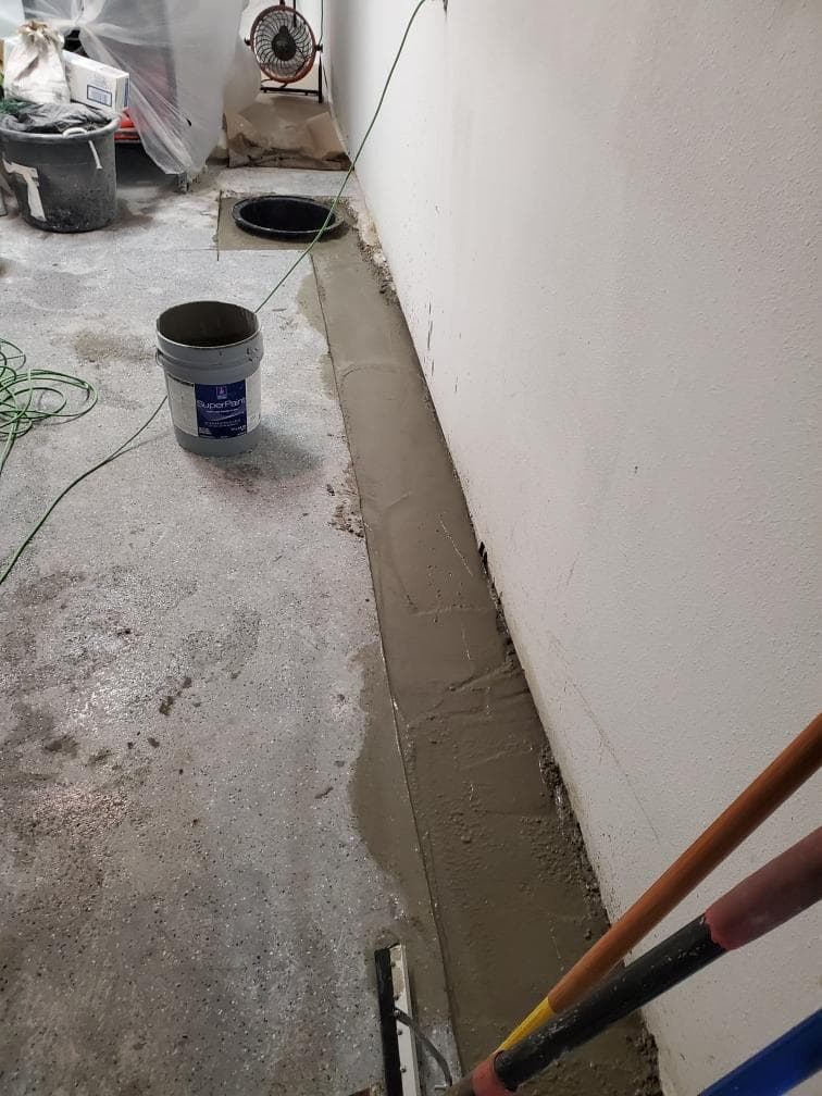 Freshly poured concrete along an interior wall covering a newly installed drainage system with a sump basin in the background Freshly poured concrete along an interior wall covering a newly installed drainage system with a sump basin in the background
