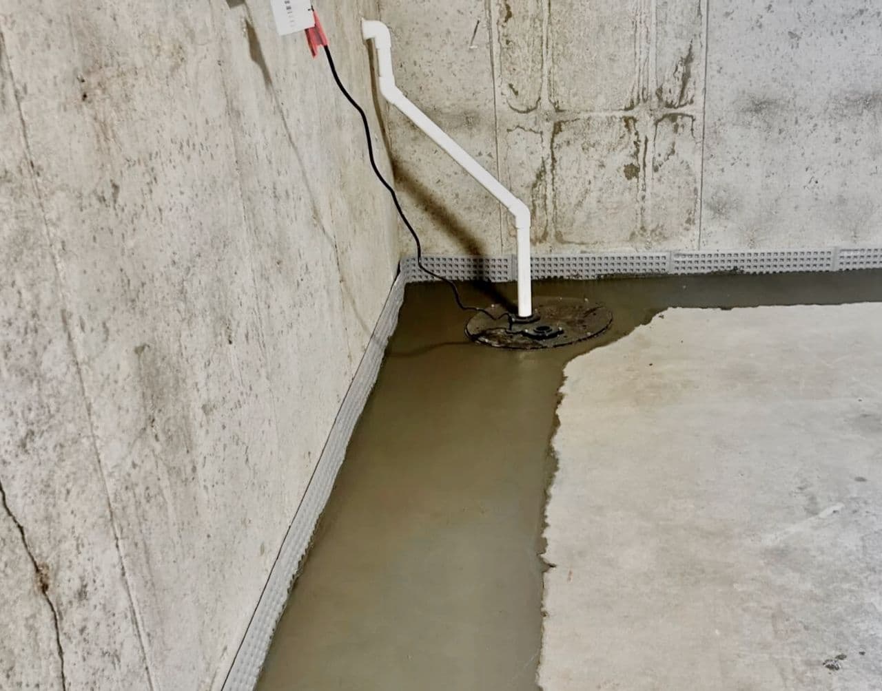 Black sump pump basin installed in the corner of a basement with a white PVC discharge pipe leading up a concrete wall Black sump pump basin installed in the corner of a basement with a white PVC discharge pipe leading up a concrete wall