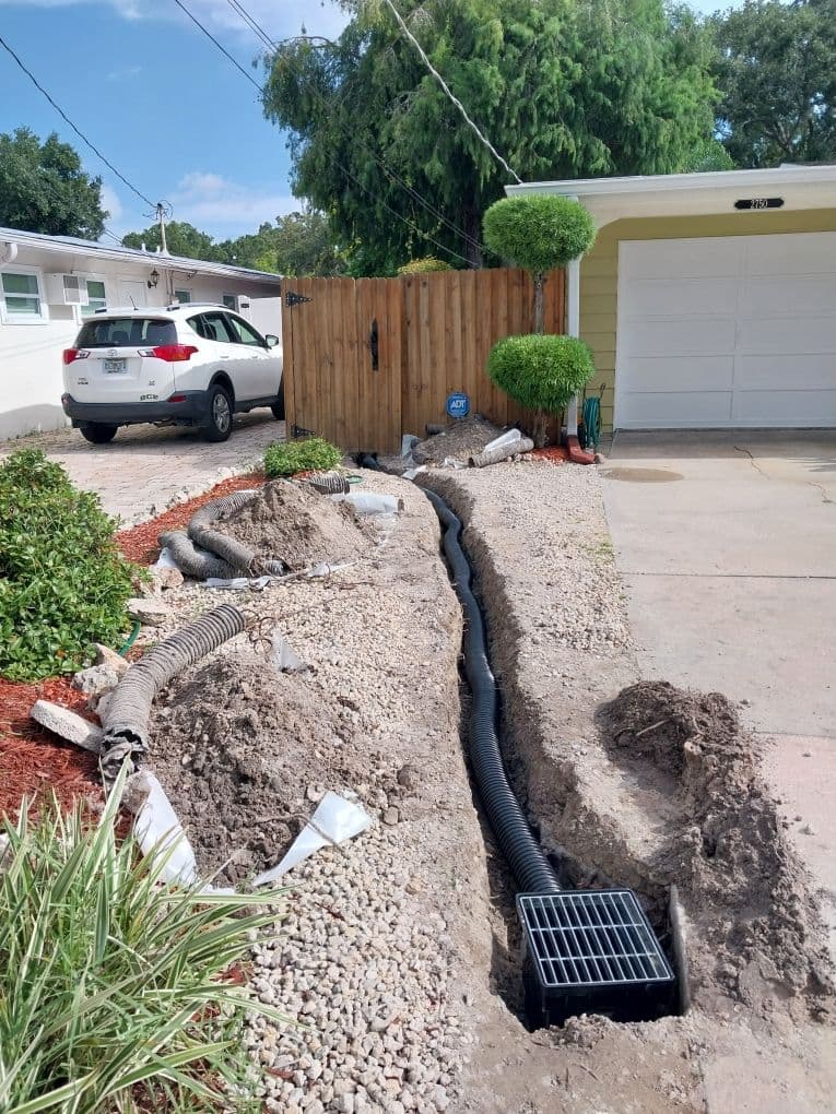 Long trench along a driveway containing a black corrugated drain pipe leading to a square catch basin grate near a wooden fences foundation Long trench along a driveway containing a black corrugated drain pipe leading to a square catch basin grate near a wooden fences foundation