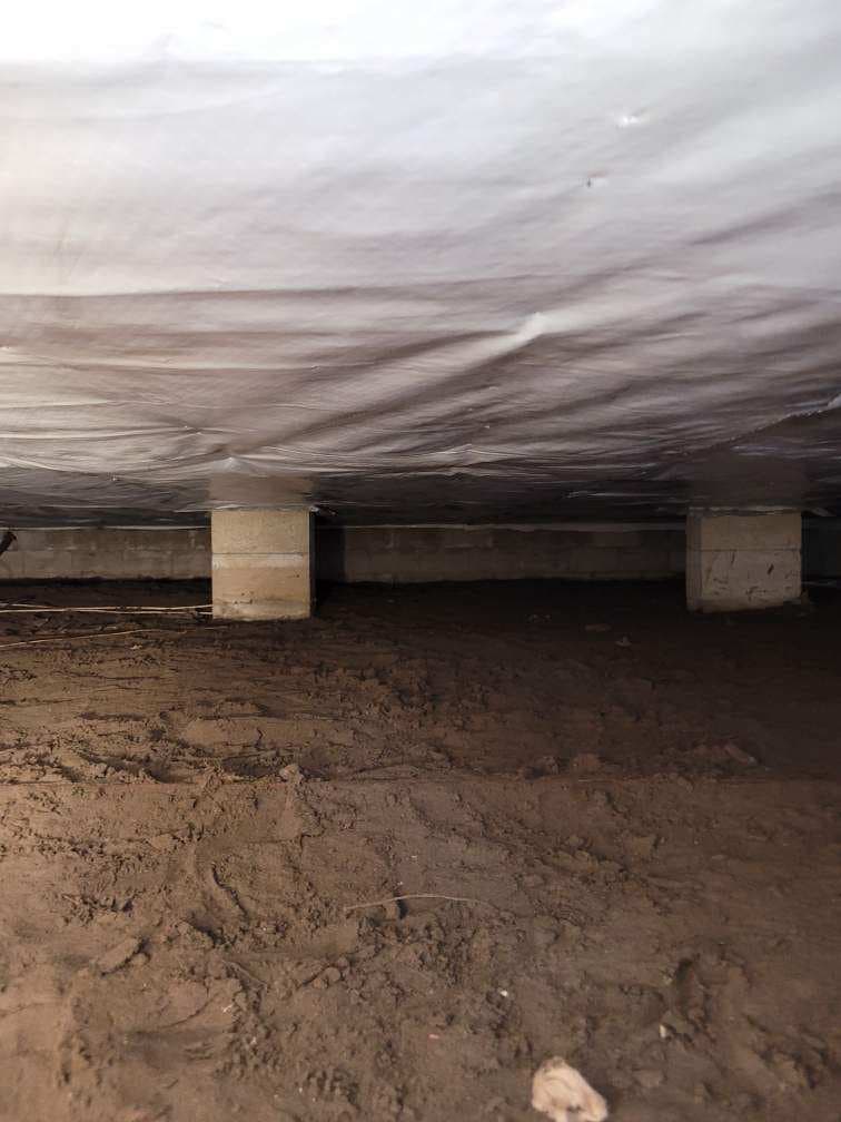 Crawlspace interior with white vapor barrier installed across floor joists, concrete block support columns and sandy soil floor visible Crawlspace interior with white vapor barrier installed across floor joists, concrete block support columns and sandy soil floor visible
