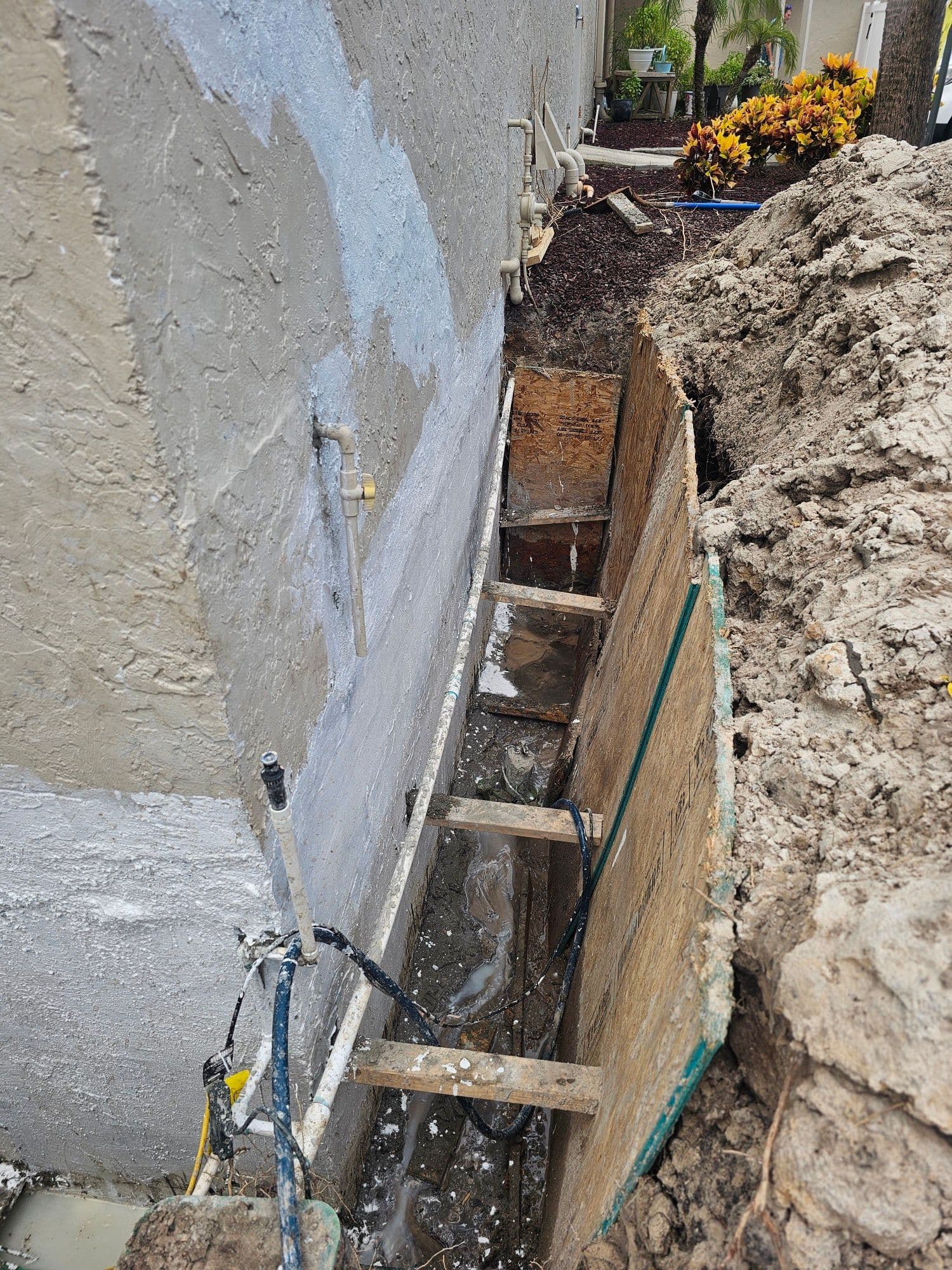 Deep exterior foundation trench next to a house wall coated in grey waterproofing material with plywood shoring and muddy water at the bottom Deep exterior foundation trench next to a house wall coated in grey waterproofing material with plywood shoring and muddy water at the bottom