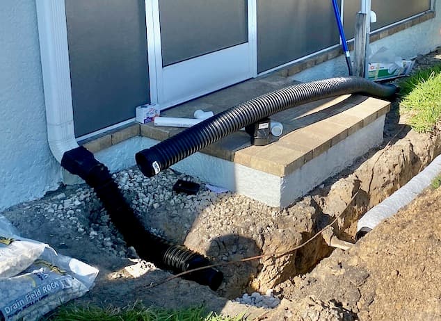 French drain corrugated pipe installation alongside a Florida home's exterior foundation with drain field rock and patio steps visible French drain corrugated pipe installation alongside a Florida home's exterior foundation with drain field rock and patio steps visible