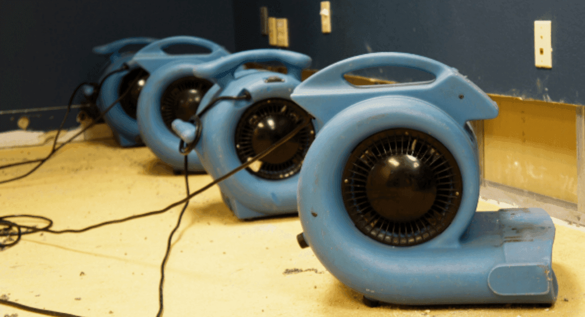 Row of industrial blue air mover fans lined up on stripped flooring during active water damage dry-out after flooding Row of industrial blue air mover fans lined up on stripped flooring during active water damage dry-out after flooding