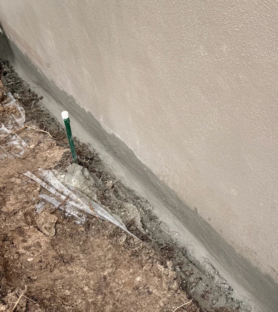 Freshly applied grey waterproofing concrete along the exterior foundation of a tan house Freshly applied grey waterproofing concrete along the exterior foundation of a tan house