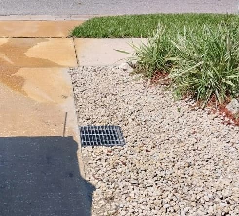 Drain grate set in a gravel bed beside a walkway and lawn, with the concrete stained where water used to run across it Drain grate set in a gravel bed beside a walkway and lawn, with the concrete stained where water used to run across it