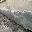 Close-up of fresh grey concrete waterproofing applied to the bottom foundation wall of a tan house Close-up of fresh grey concrete waterproofing applied to the bottom foundation wall of a tan house